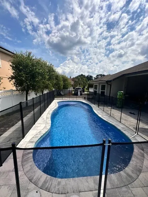 Narrow backyard pool with blue water, surrounded by fencing and houses under a cloudy sky