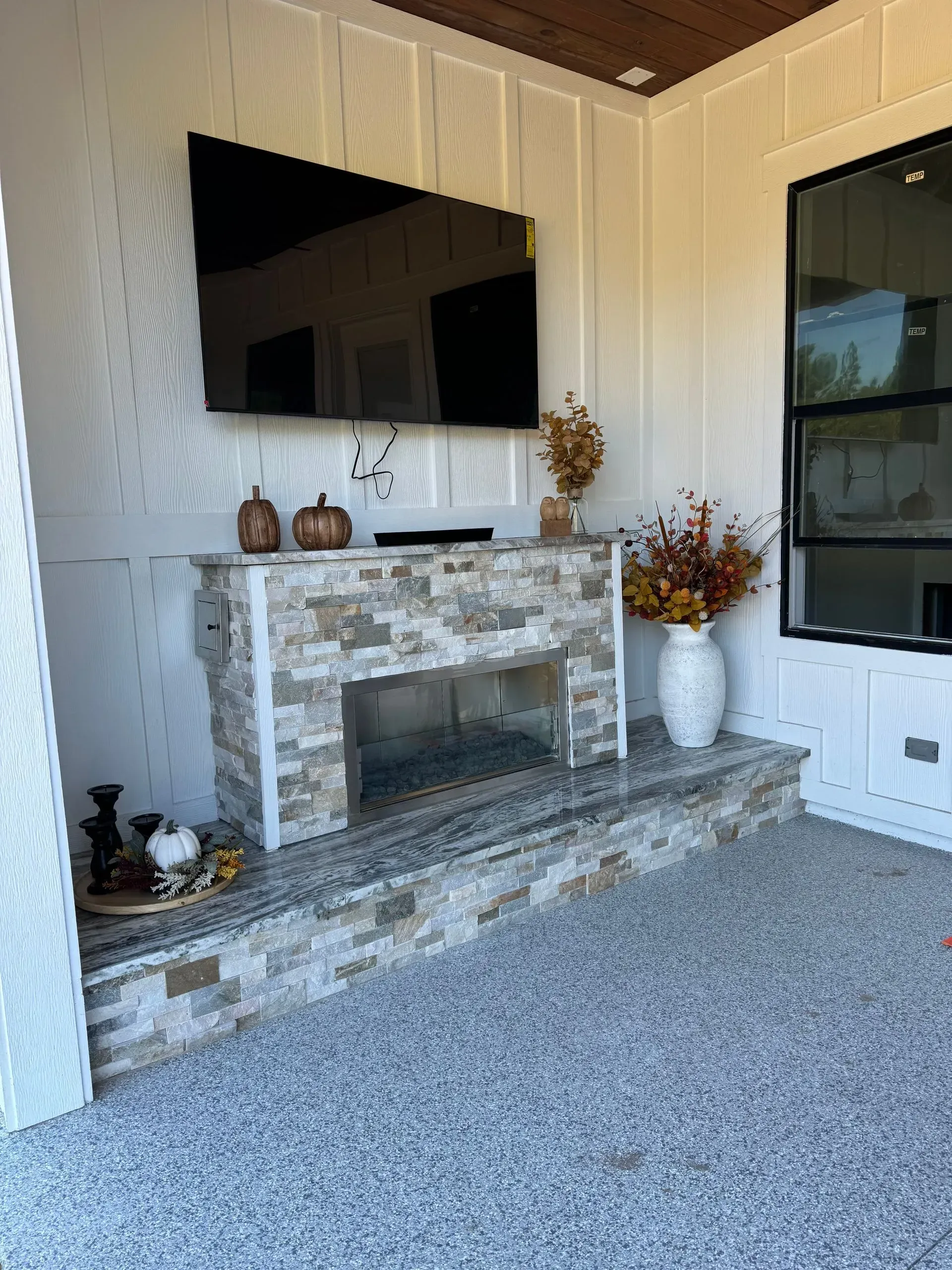 Living room with wall-mounted TV, stone fireplace, and white paneled walls with autumn decor