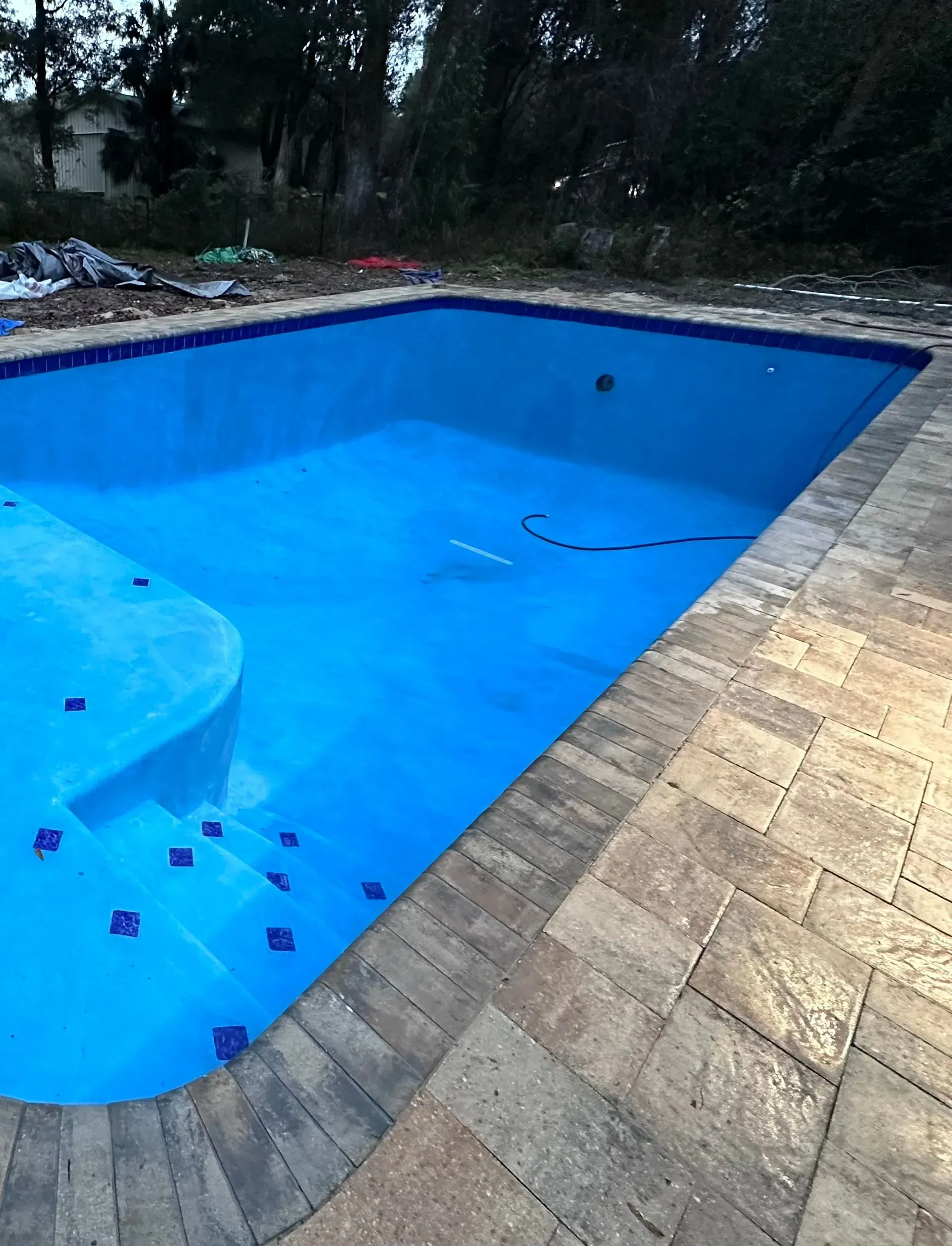 Empty blue swimming pool with tan stone deck and trees in the background