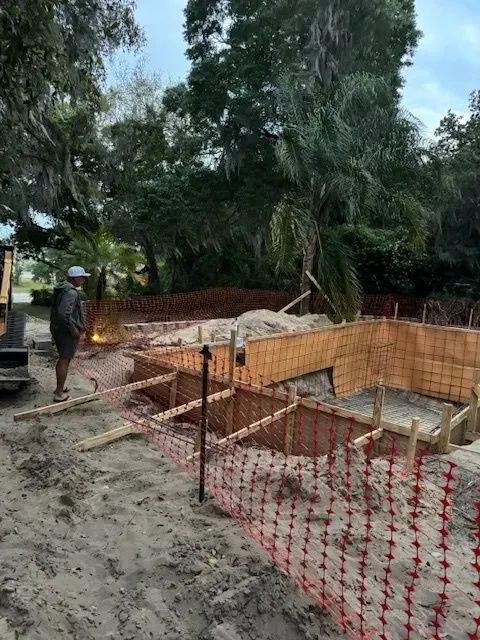 Construction site with foundation forms, red safety netting, and a worker near trees on a muddy lot