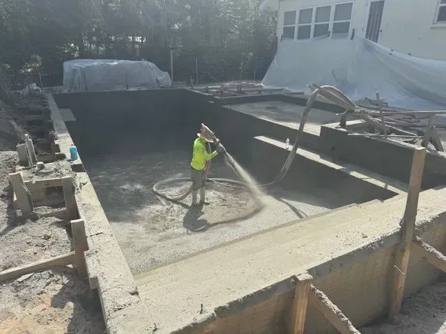 Worker pressure-washing a concrete pool under construction in a sunlit backyard