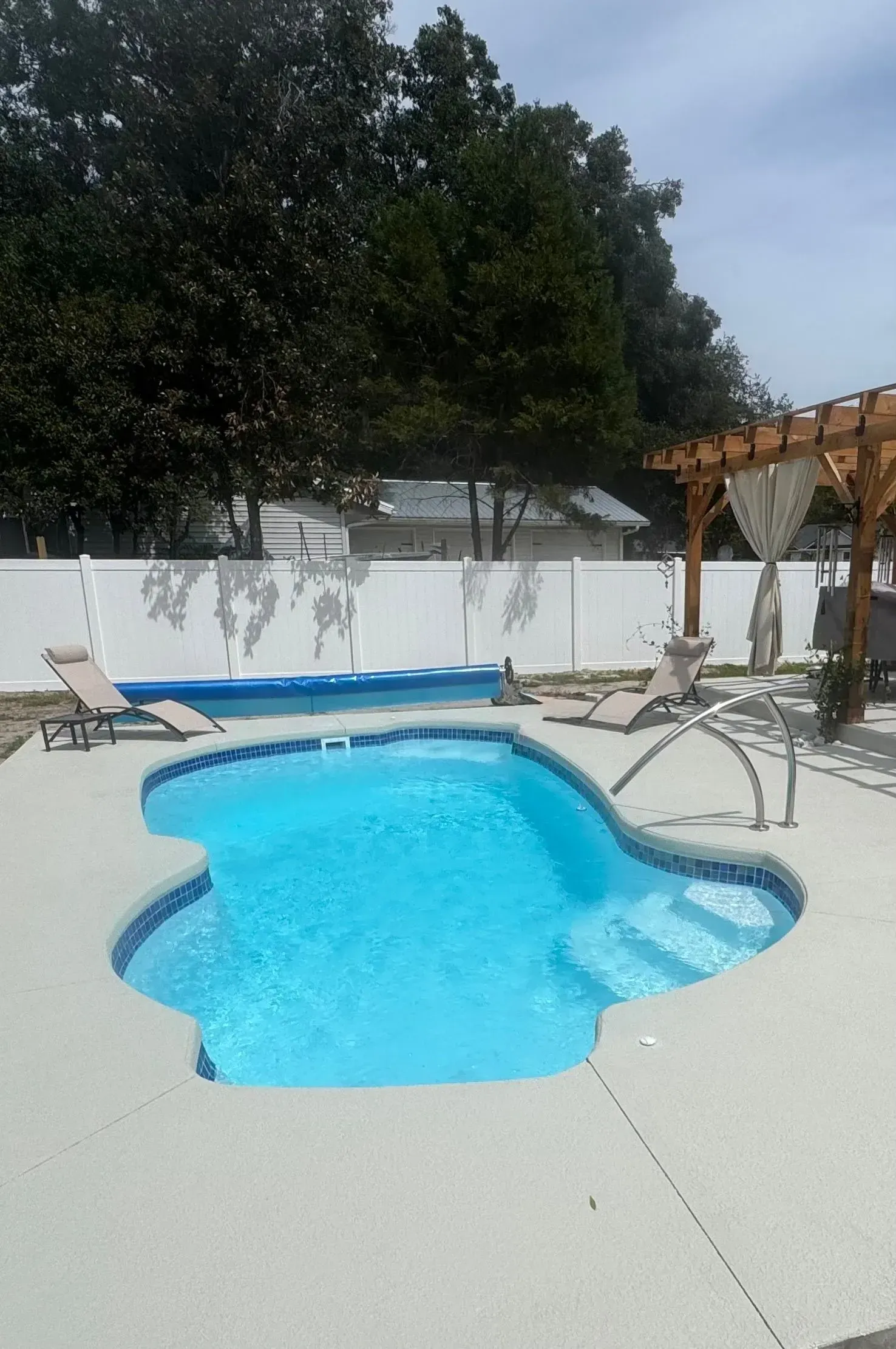 Backyard pool with bright blue water, white deck, lounge chairs, and wooden pergola under trees