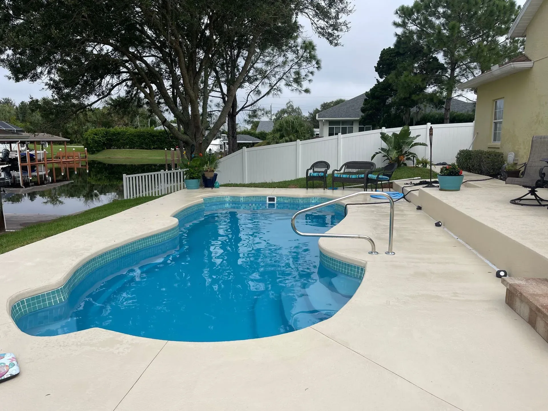Backyard swimming pool with blue water, patio chairs, and a white fence on a cloudy day
