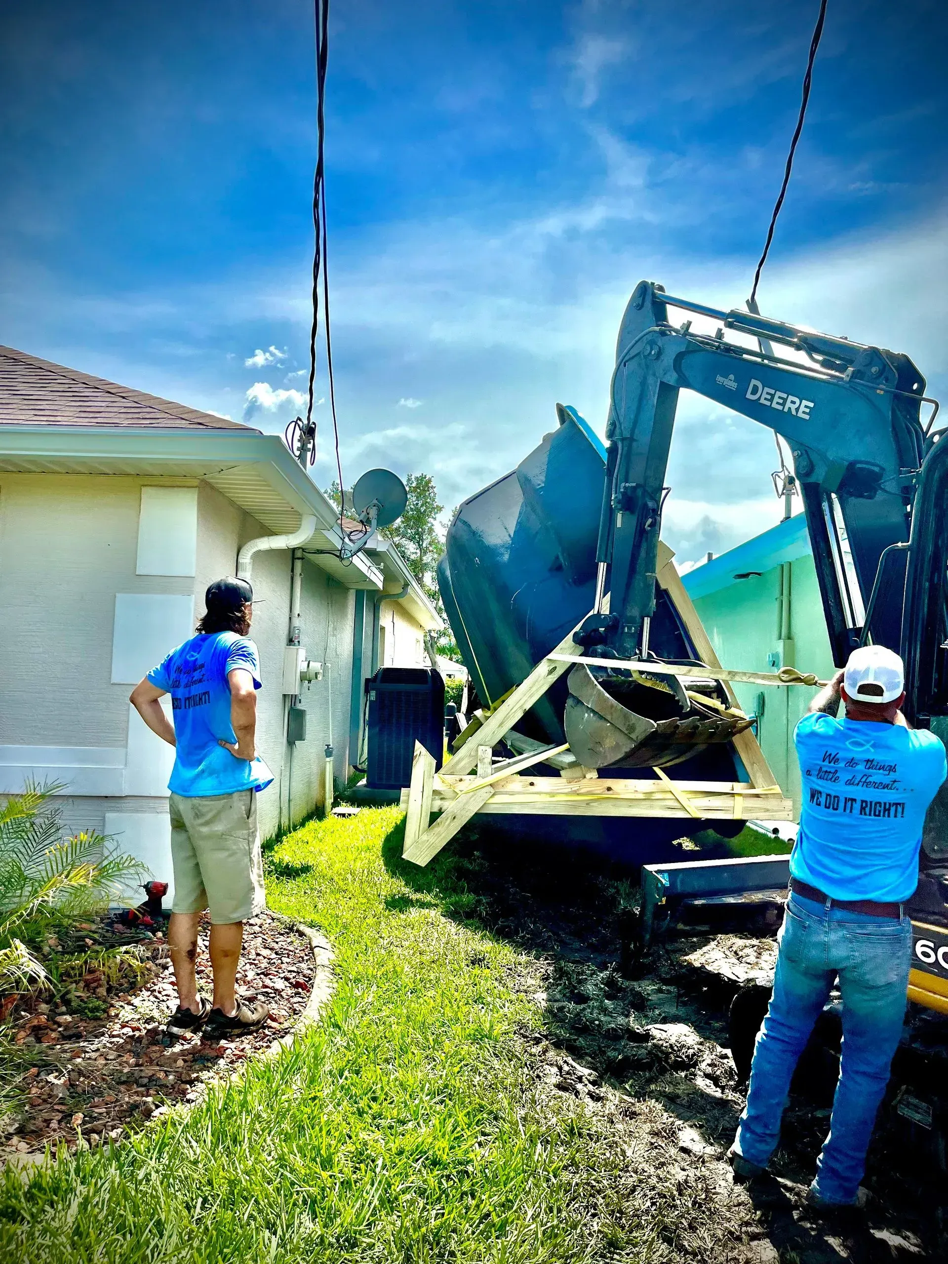 Workers load debris into a blue excavator beside a house under a sunny sky