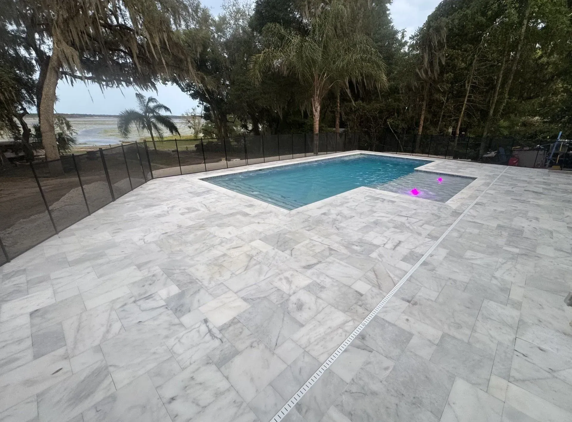 White stone patio beside a rectangular swimming pool, surrounded by trees and a fence.
