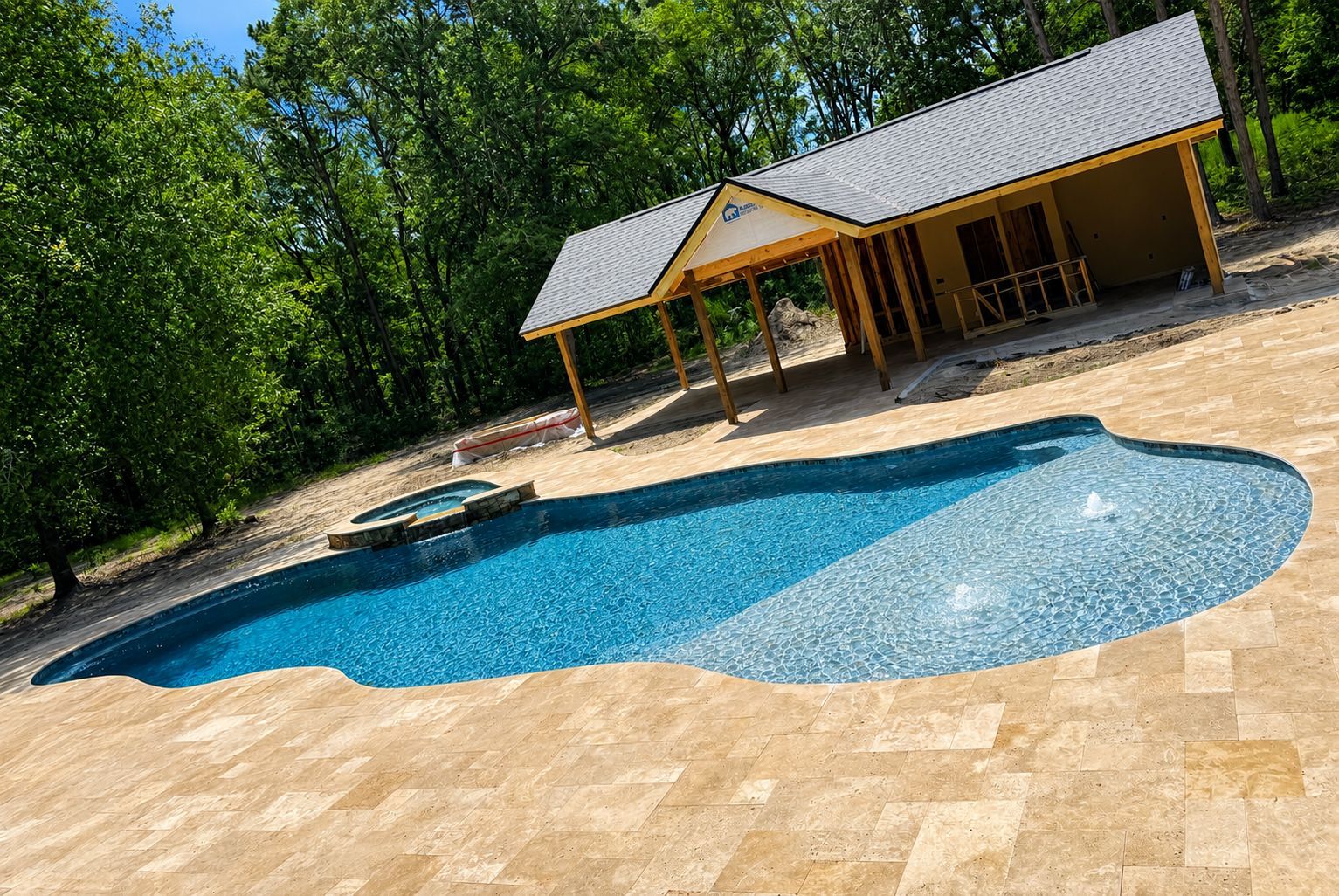 Curved blue swimming pool beside a tan stone deck and wooden pavilion near trees