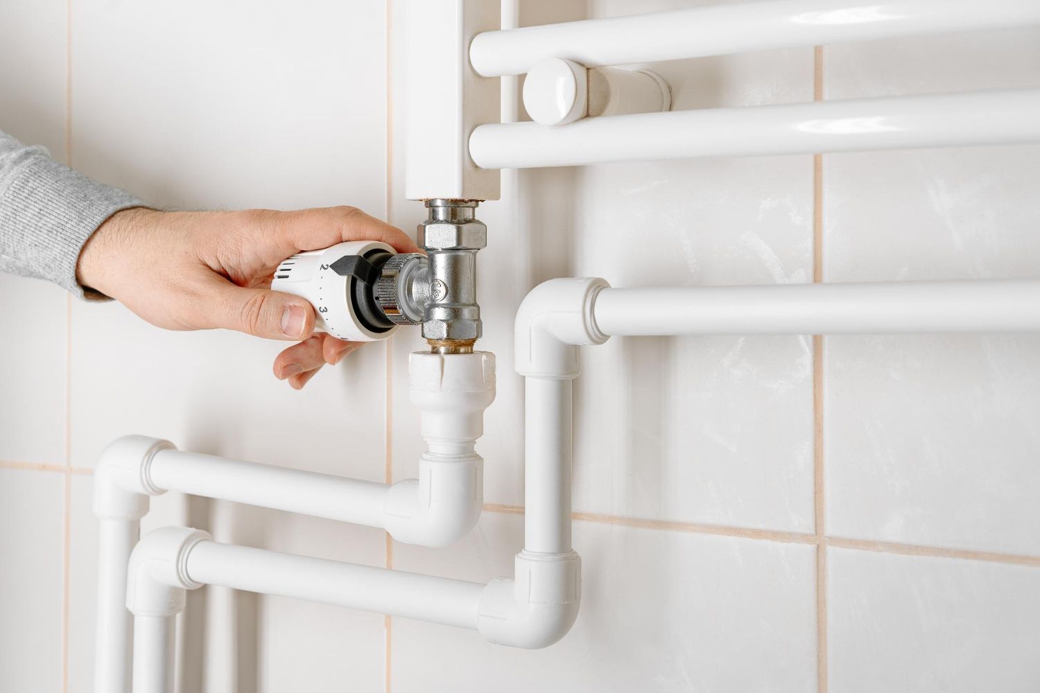 Hand adjusting a radiator valve on white pipes against a tiled wall