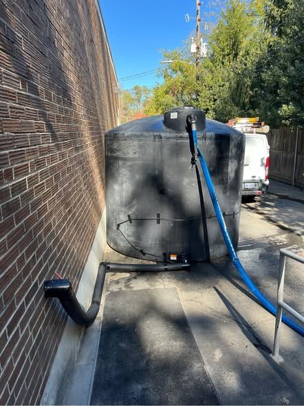 Blue hose connected to a large black tank beside a brick wall on a sunny sidewalk