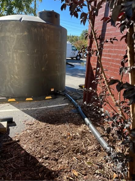 Large tan water tank beside a brick wall, with a metal pipe and dry mulch in a narrow outdoor service area