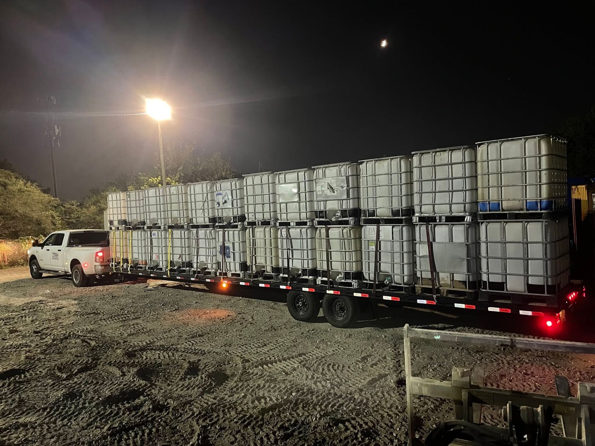 White truck hauling stacked industrial containers on a flatbed trailer at night under a bright streetlight.