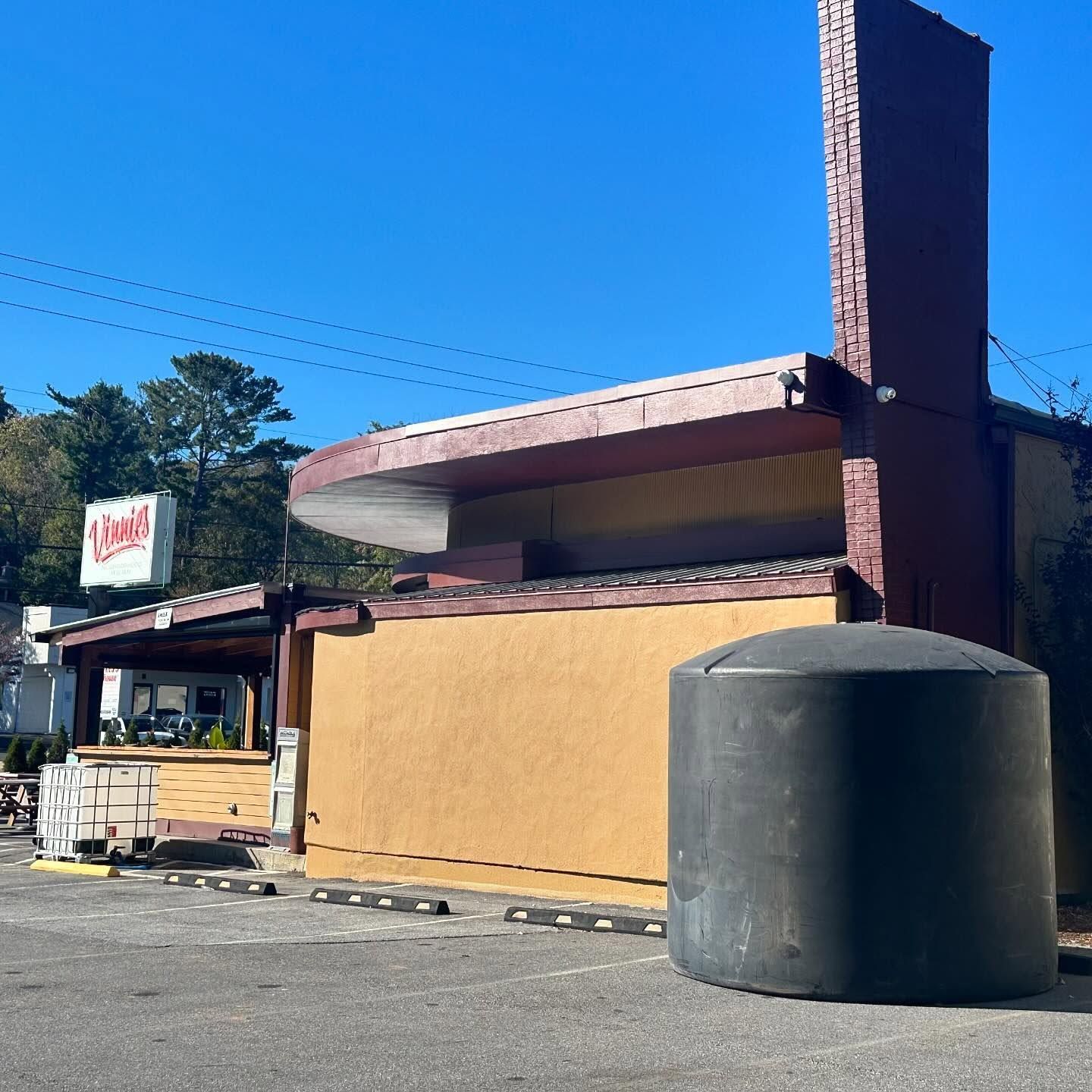 Small roadside building with tan walls, red roof, and a large black water tank in front under a clear blue sky