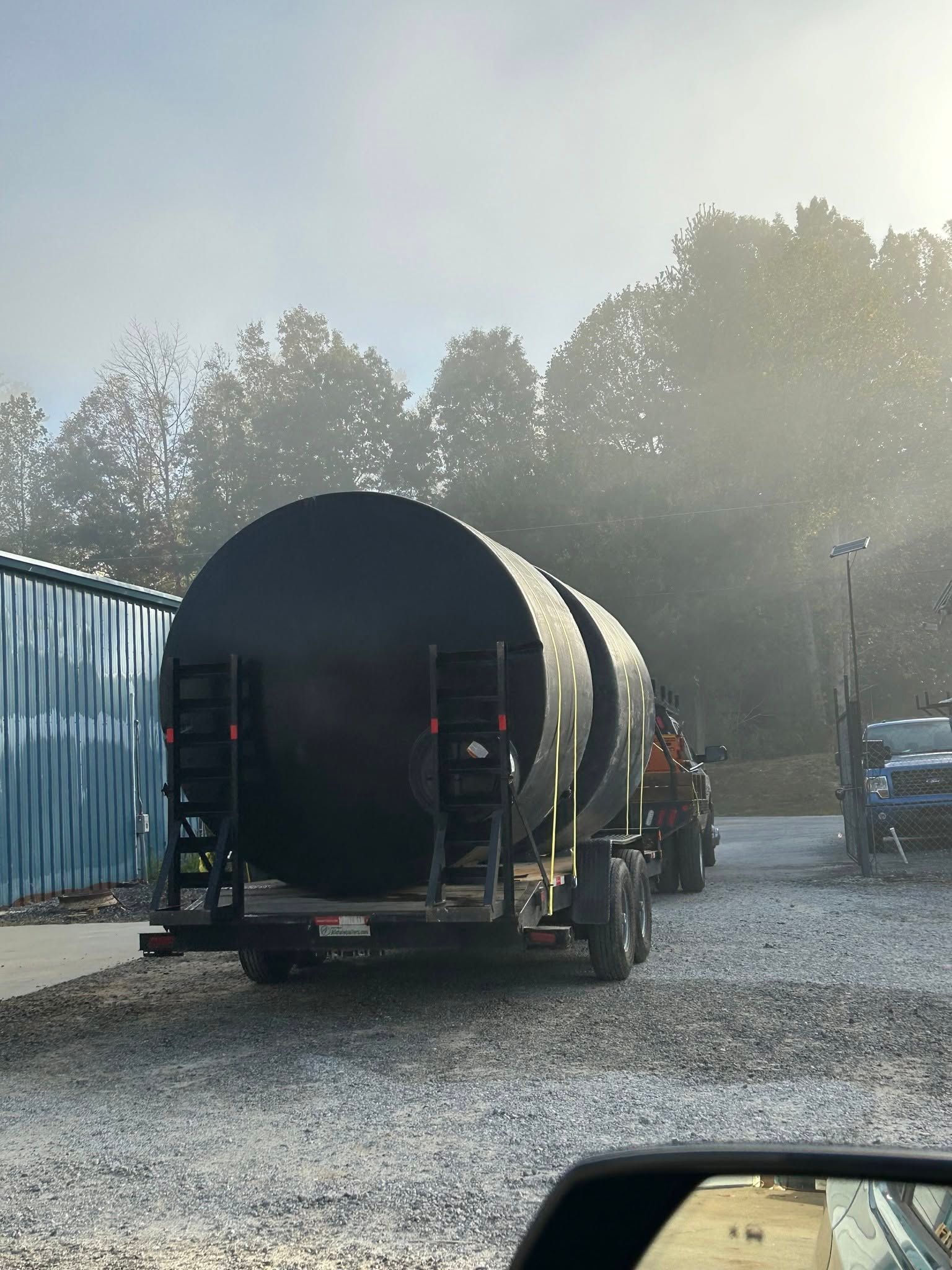Large black cylindrical tank on a trailer in a gravel yard, with a blue container and trees in the background.