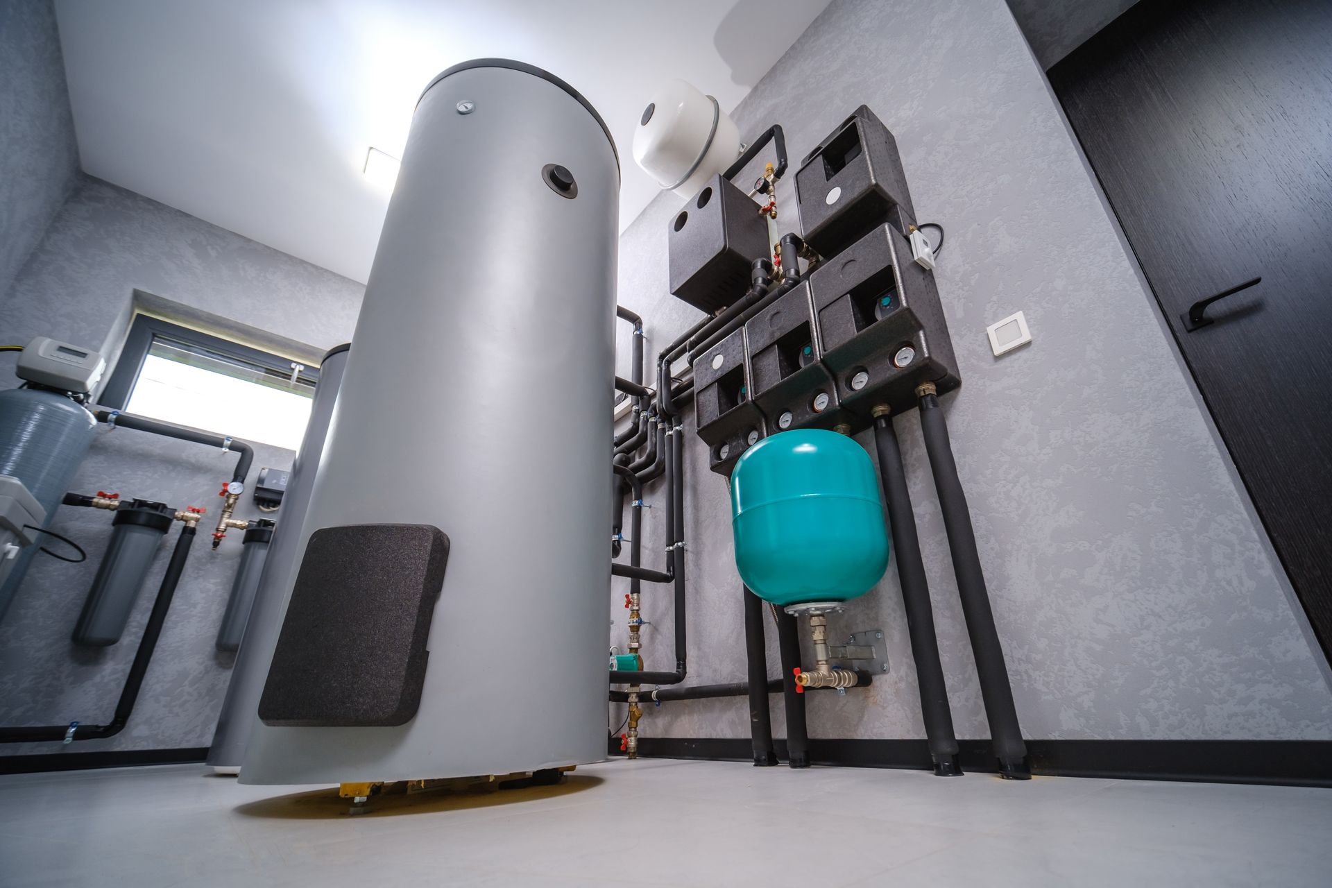 Indoor boiler and water tank setup in a utility room with pipes, valves, and a gray cylindrical unit