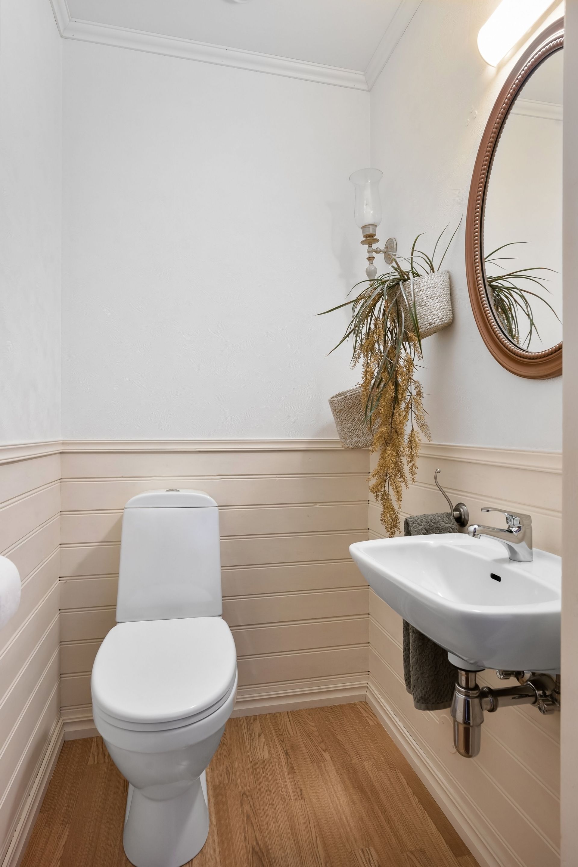 Small bathroom with white toilet, wall-mounted sink, round mirror, and dried plant decor.