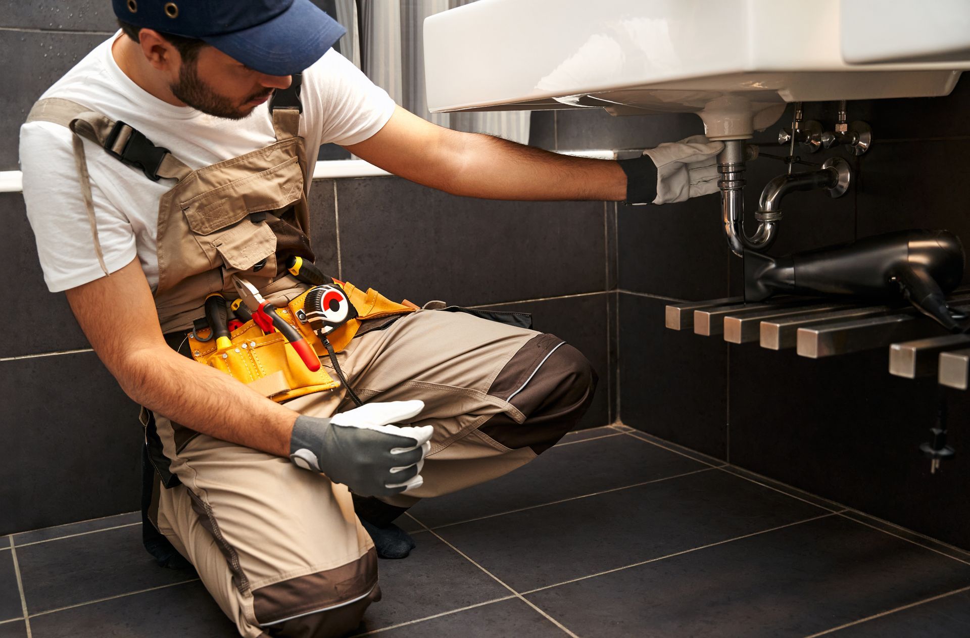 Technician kneeling under a sink, inspecting plumbing with tools in a tiled kitchen.