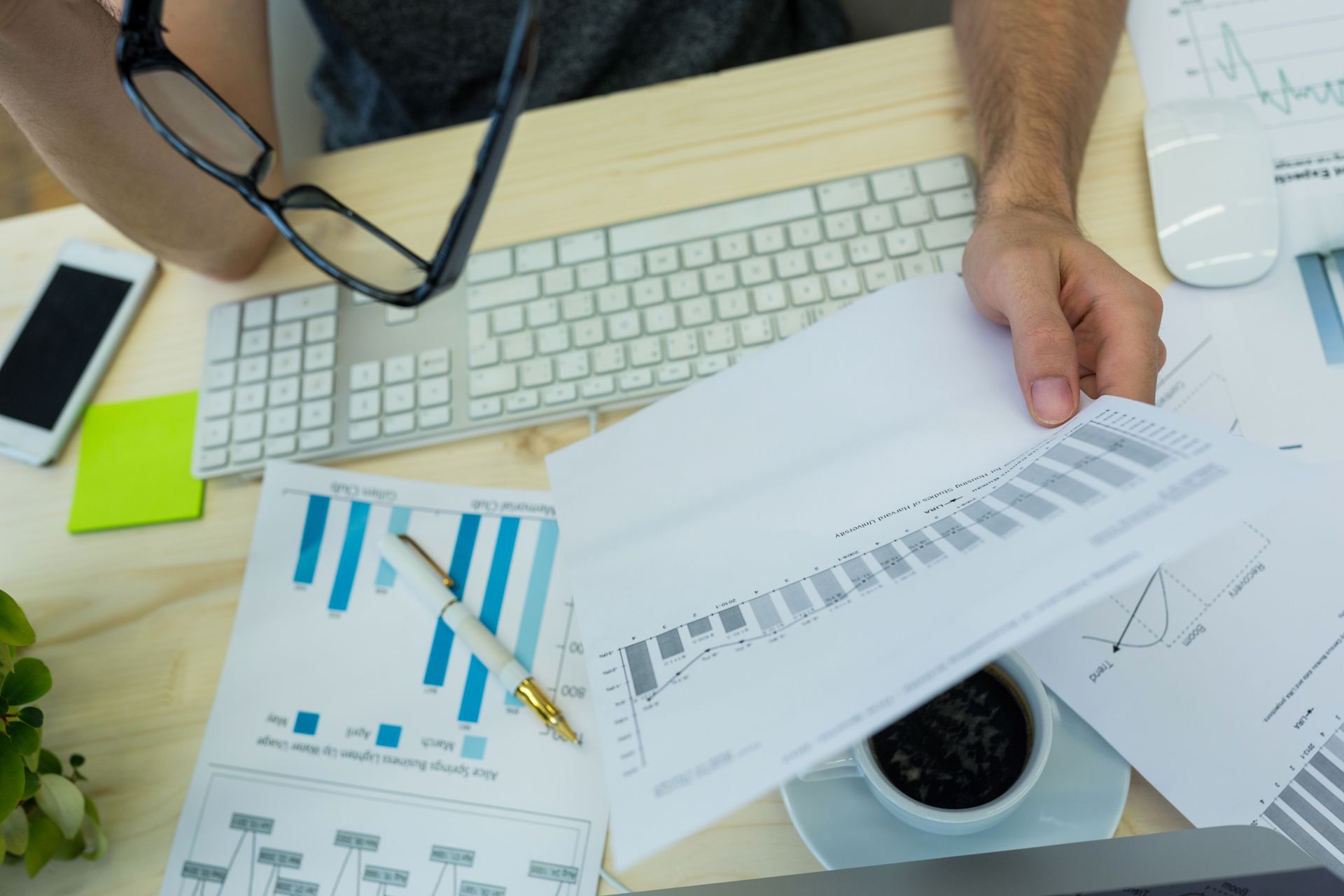 Hands reviewing business charts and reports beside a keyboard, phone, and coffee cup