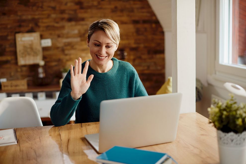 Woman waving during a video call at a laptop in a bright home office