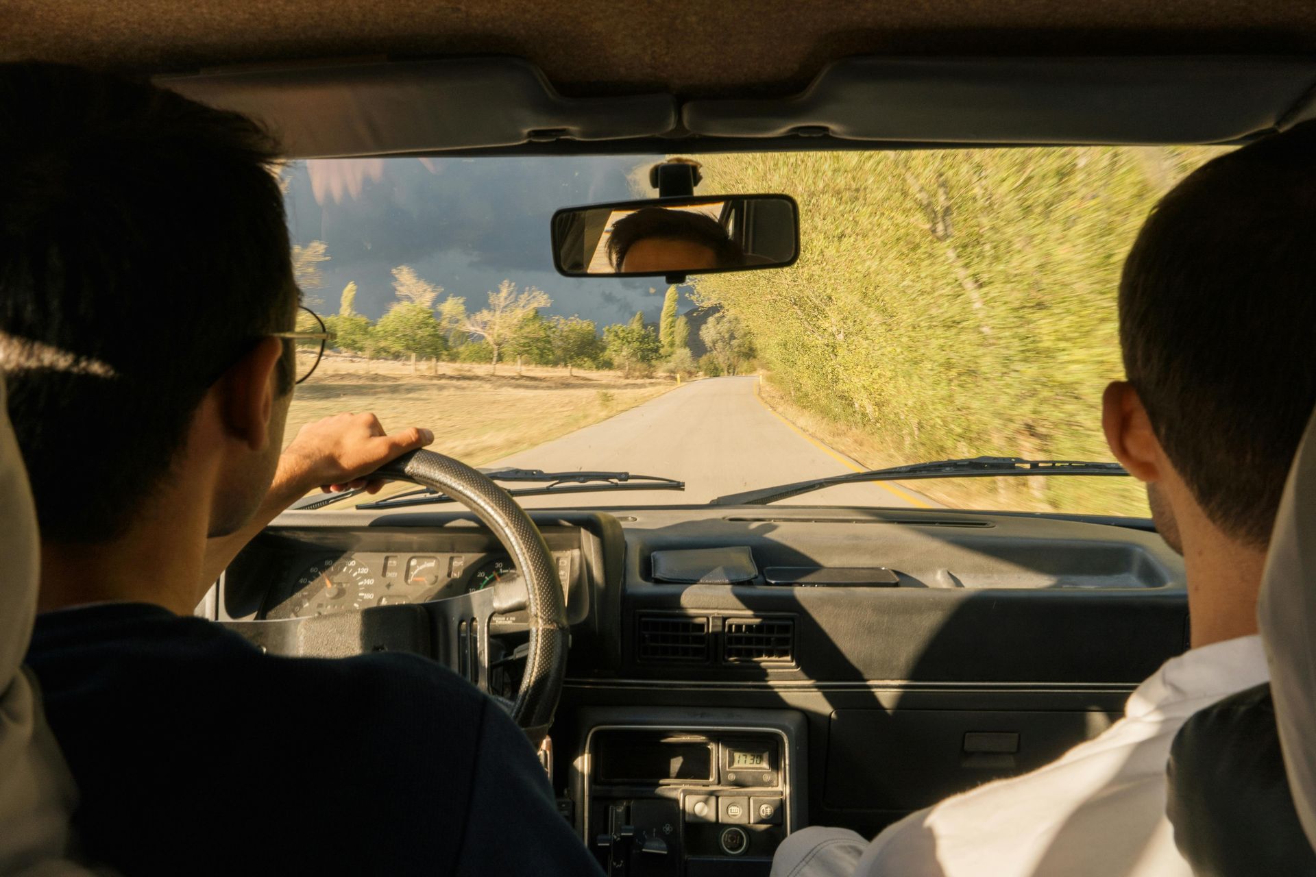 Two people in a car driving on a sunlit dirt road, with one pointing ahead.