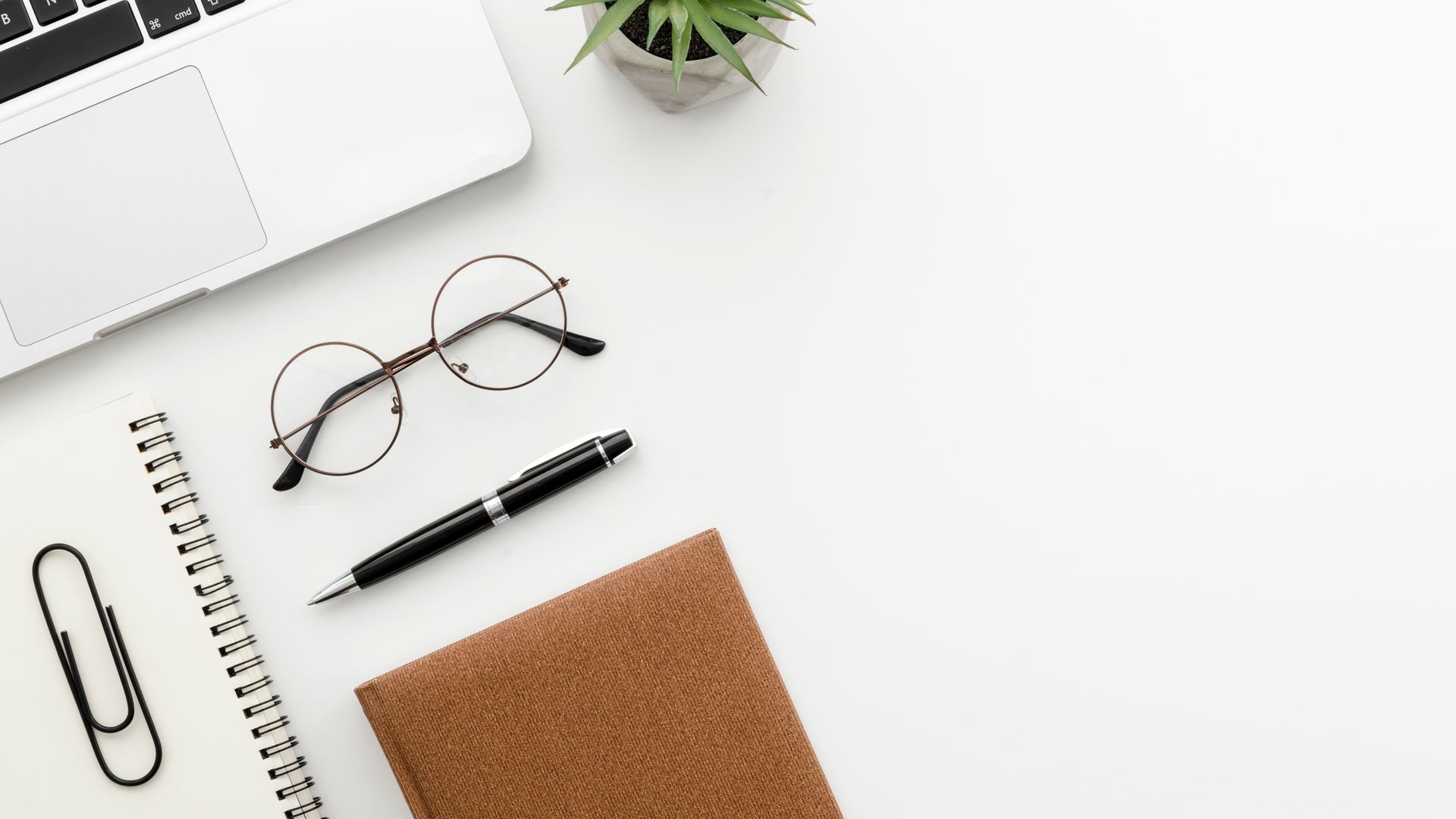 Flat lay of a white desk with laptop, glasses, pen, notebook, paperclip, and a small plant