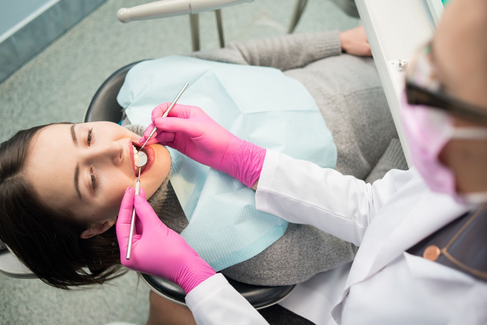 Female emergency dentist with pink gloves checks teeth of female patient sitting on dental chair.