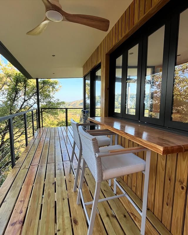 Covered wooden deck with bar seating, ceiling fan, and glass doors overlooking trees and distant hills