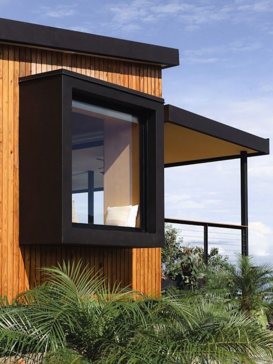 Modern tiny house with large window, wooden exterior, and covered porch overlooking greenery