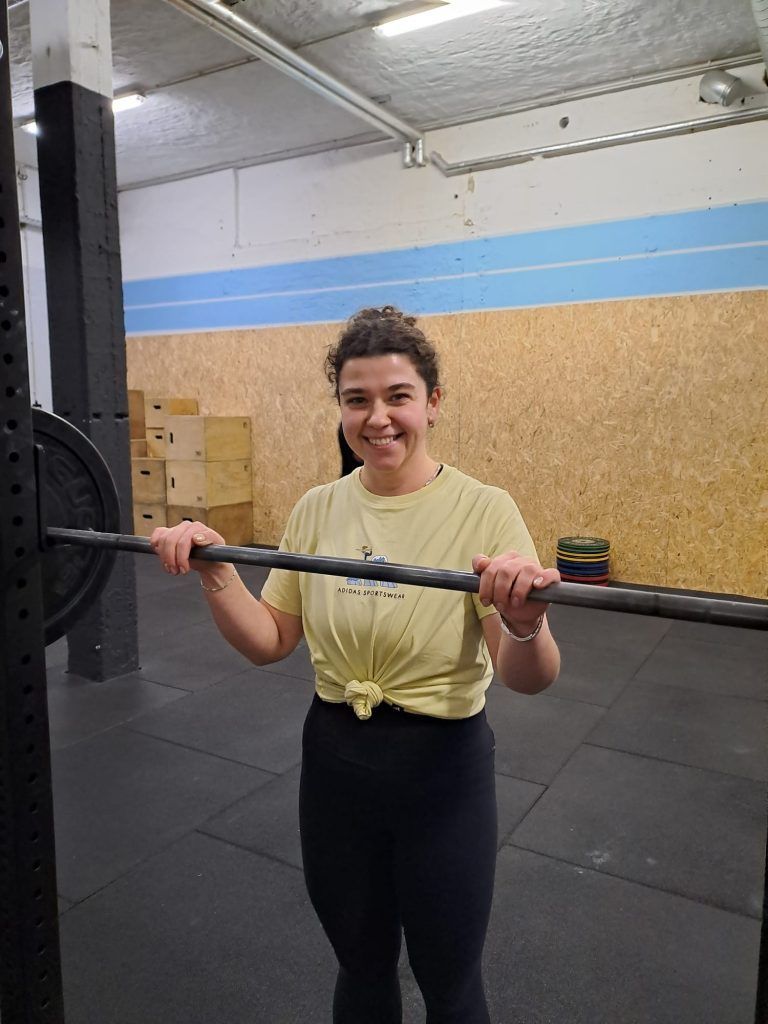 Smiling person in a yellow T-shirt standing in a gym, holding onto a barbell mounted on a squat rack.