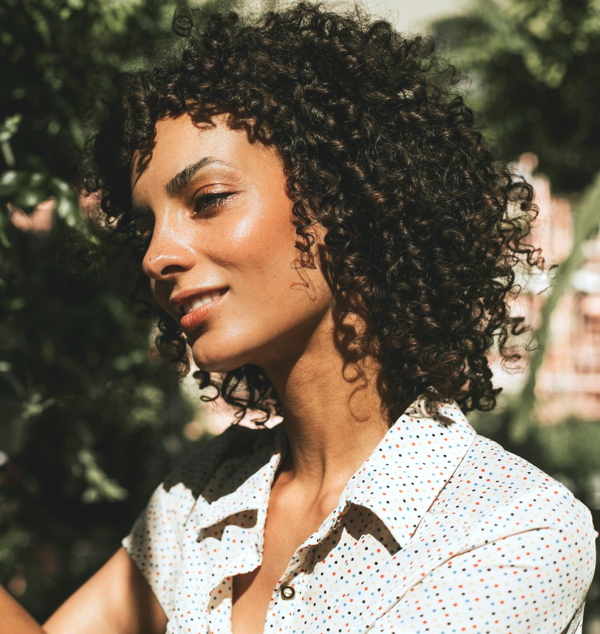 Curly-haired person in a white polka-dot shirt, smiling outdoors in dappled sunlight.