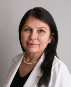 Portrait of a woman with long dark hair in a white coat and pearl necklace, against a neutral background
