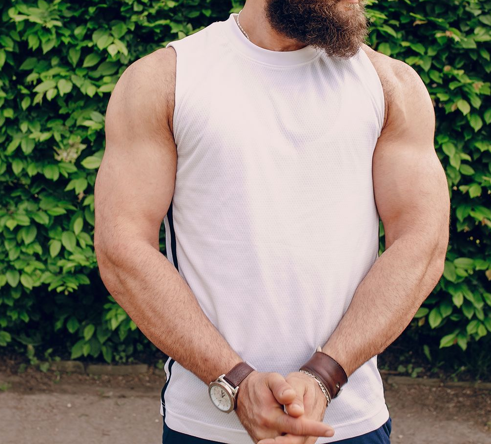 Man in a white sleeveless shirt standing with hands clasped in front of a leafy wall