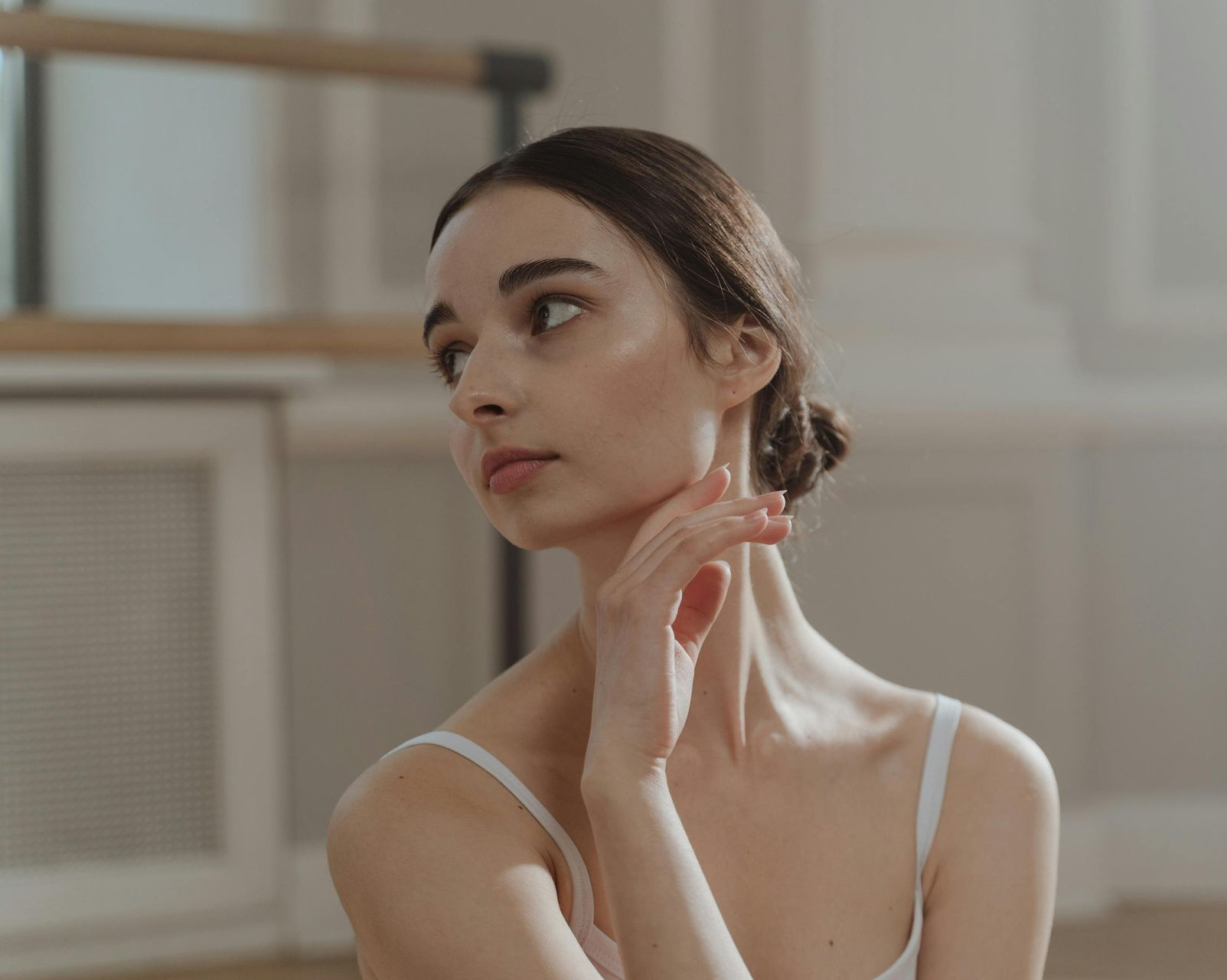 Woman in a white tank top touching her neck, looking aside in a softly lit room.