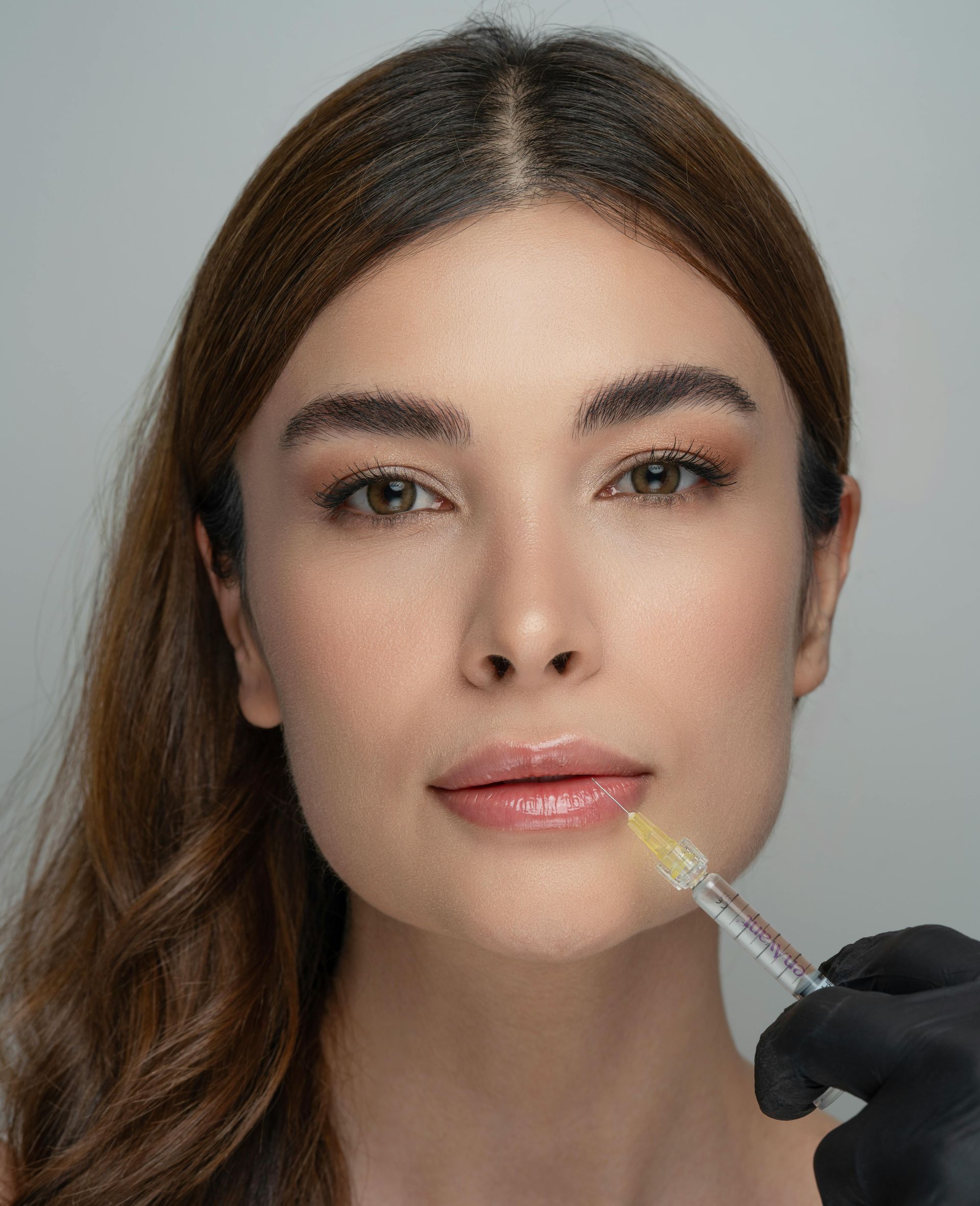 Woman with neutral makeup while a gloved hand applies lip gloss with a wand