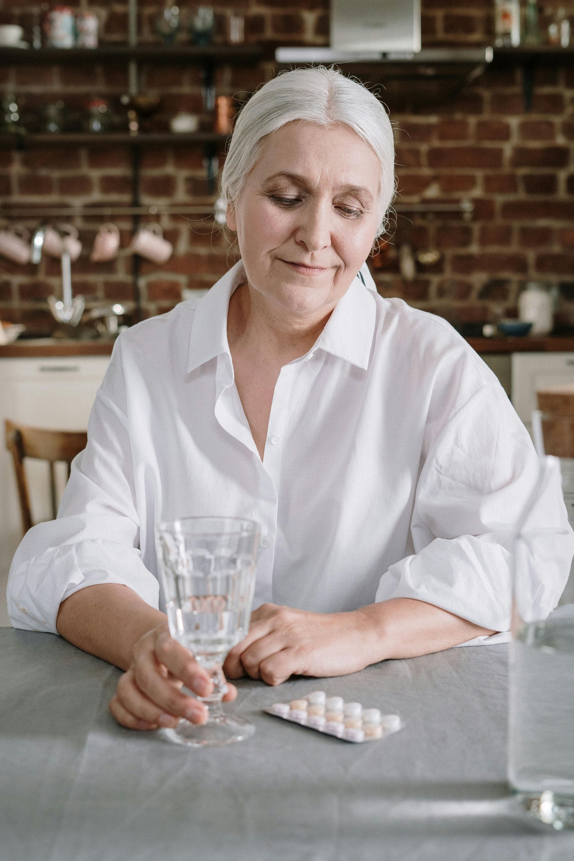 Woman in a white shirt sitting at a table with a glass of water and a blister pack of pills.
