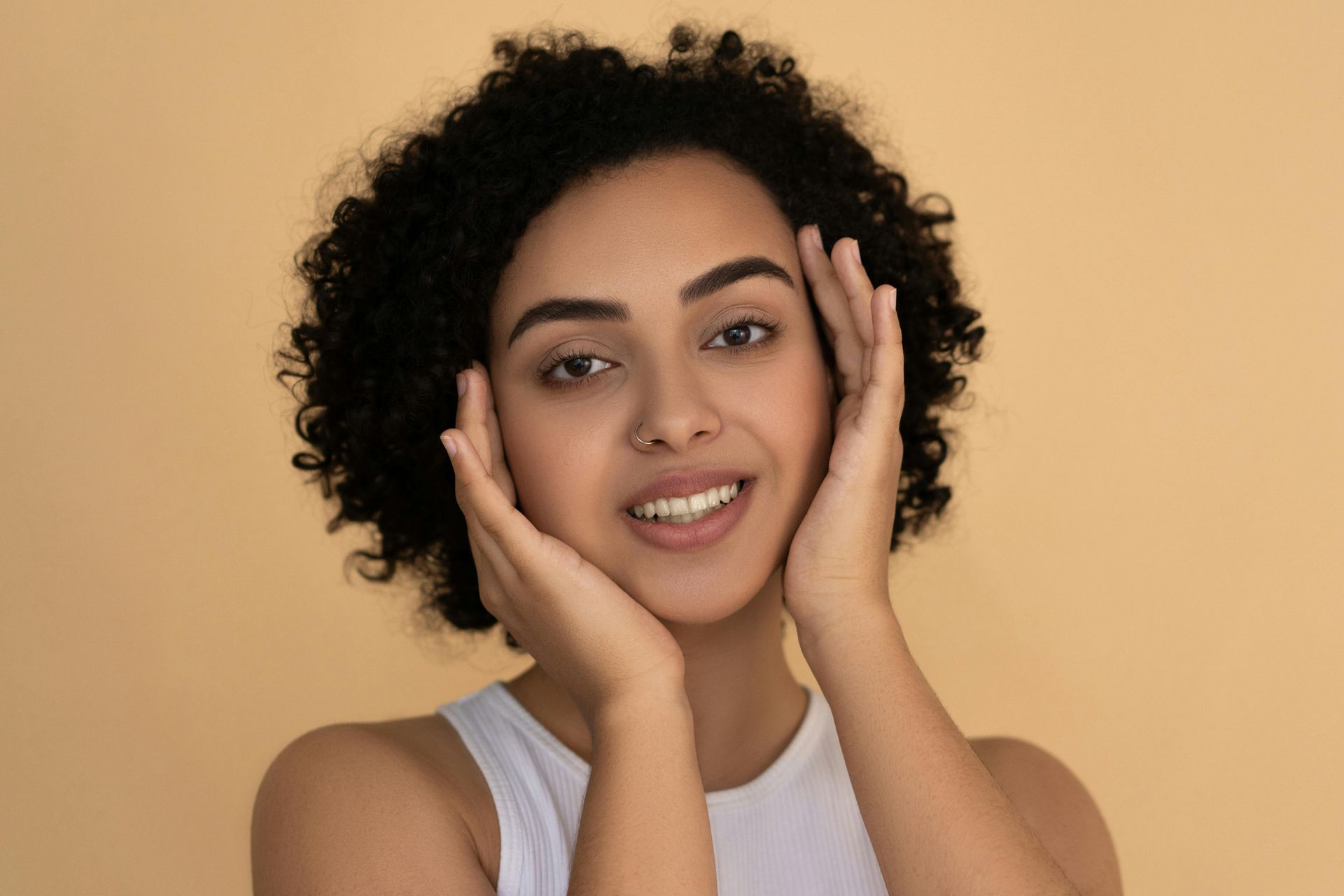 Smiling person with curly hair gently touching their face against a beige background