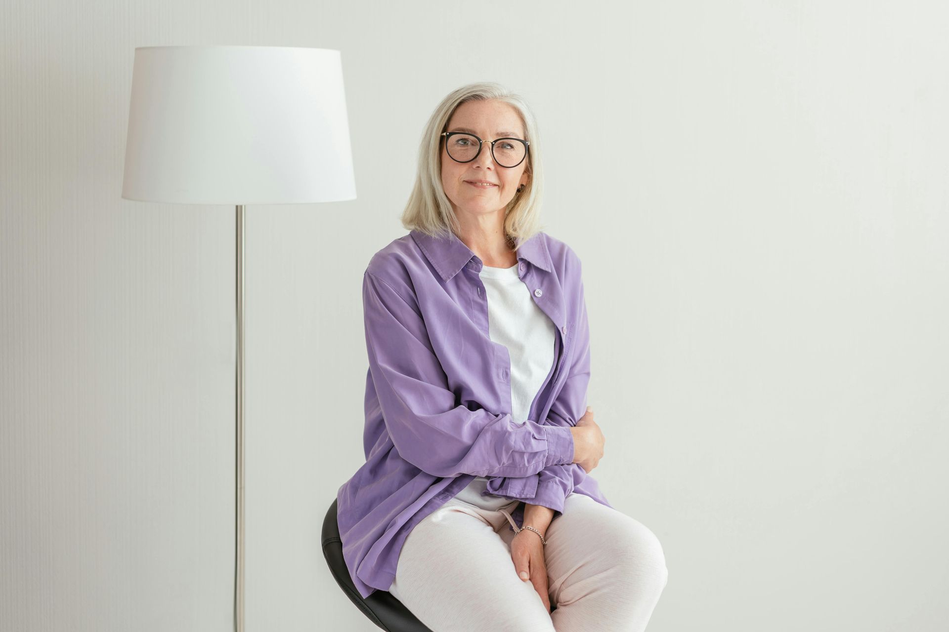 Woman in a purple shirt and white pants seated beside a floor lamp against a light wall