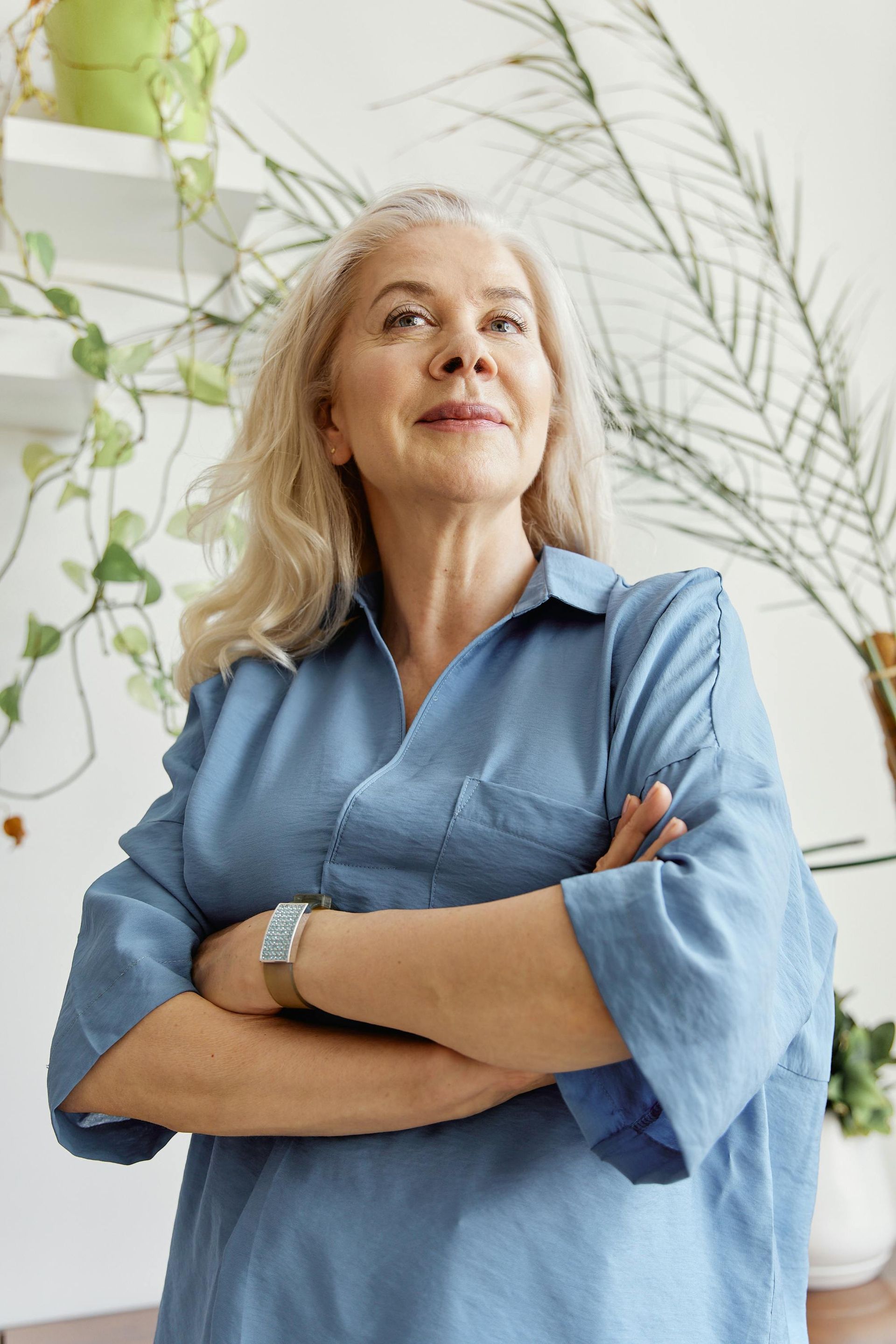 Smiling woman in a blue shirt standing with arms crossed in a bright room with plants