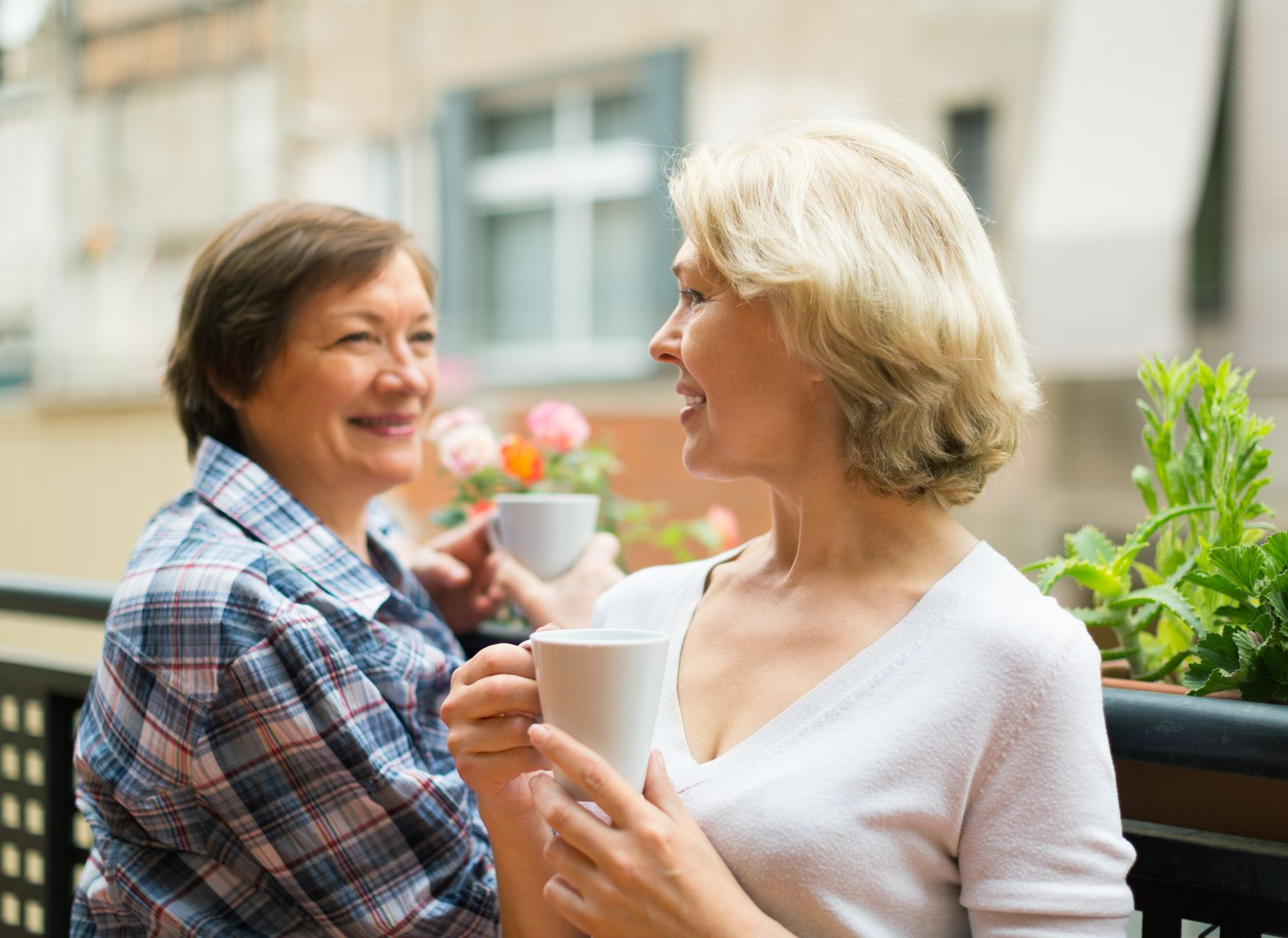 Two people chatting on a balcony, smiling and holding mugs with potted plants nearby
