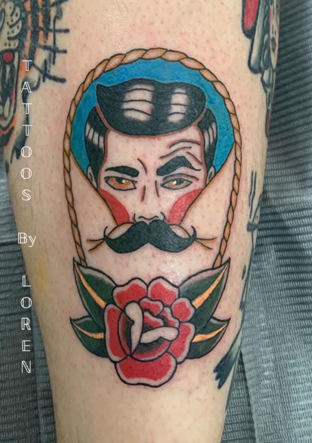 Tattoo of a man with a mustache in a rope frame above a red rose. Blue, black, green and red colors.