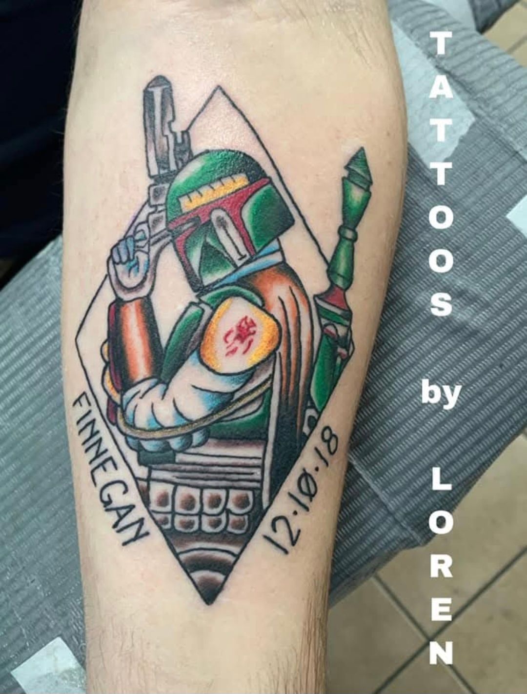 Tattoo of Boba Fett in diamond, holding weapons. Green, orange, black ink, with date and name on arm.