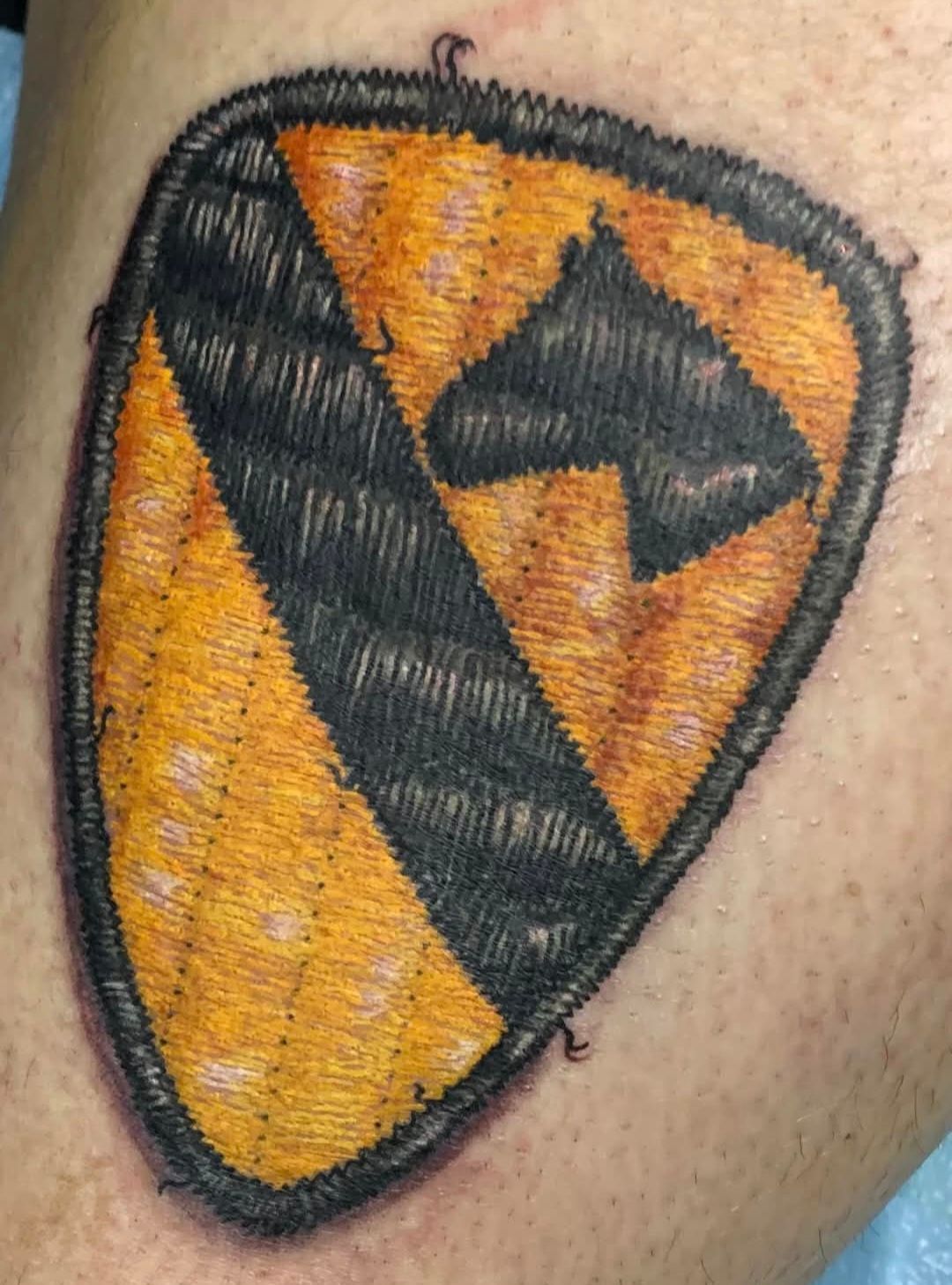 Tattoo of the U.S. Army 1st Cavalry Division patch on skin: yellow and black shield with a diagonal stripe and horse head.