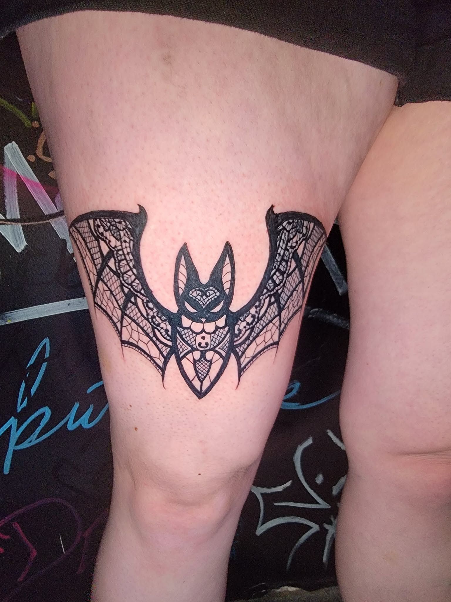 Bat tattoo on thigh, black lace-like design.