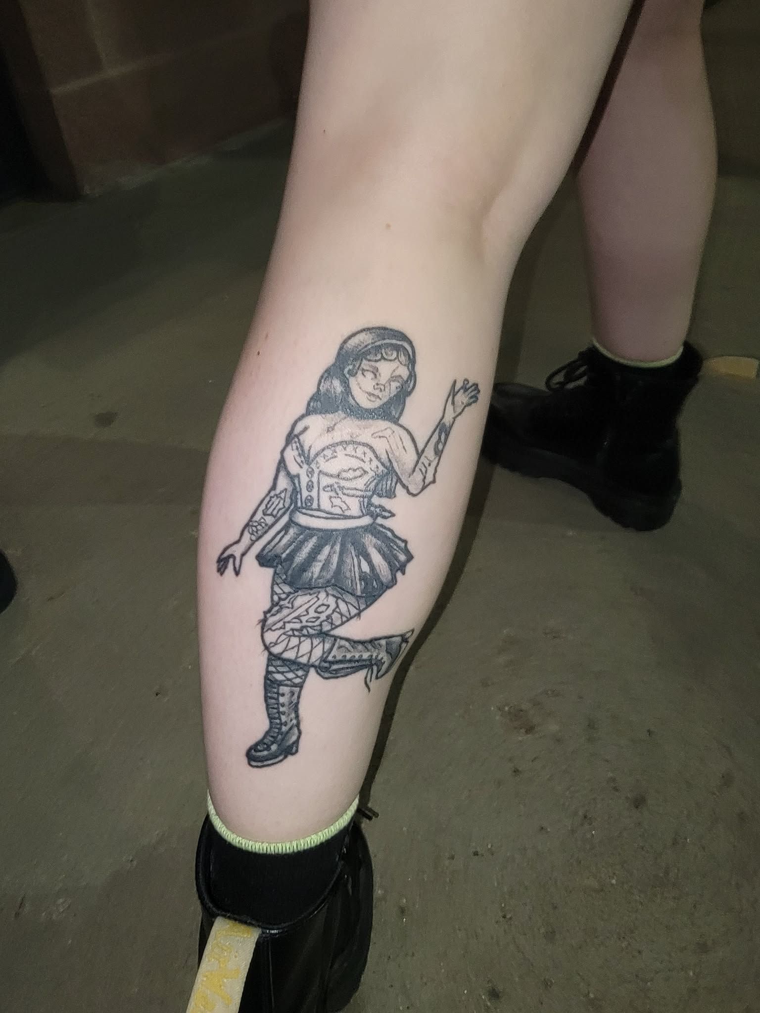 Tattoo of a dancing woman on a leg, wearing a corset and skirt. Person is wearing black boots and socks.