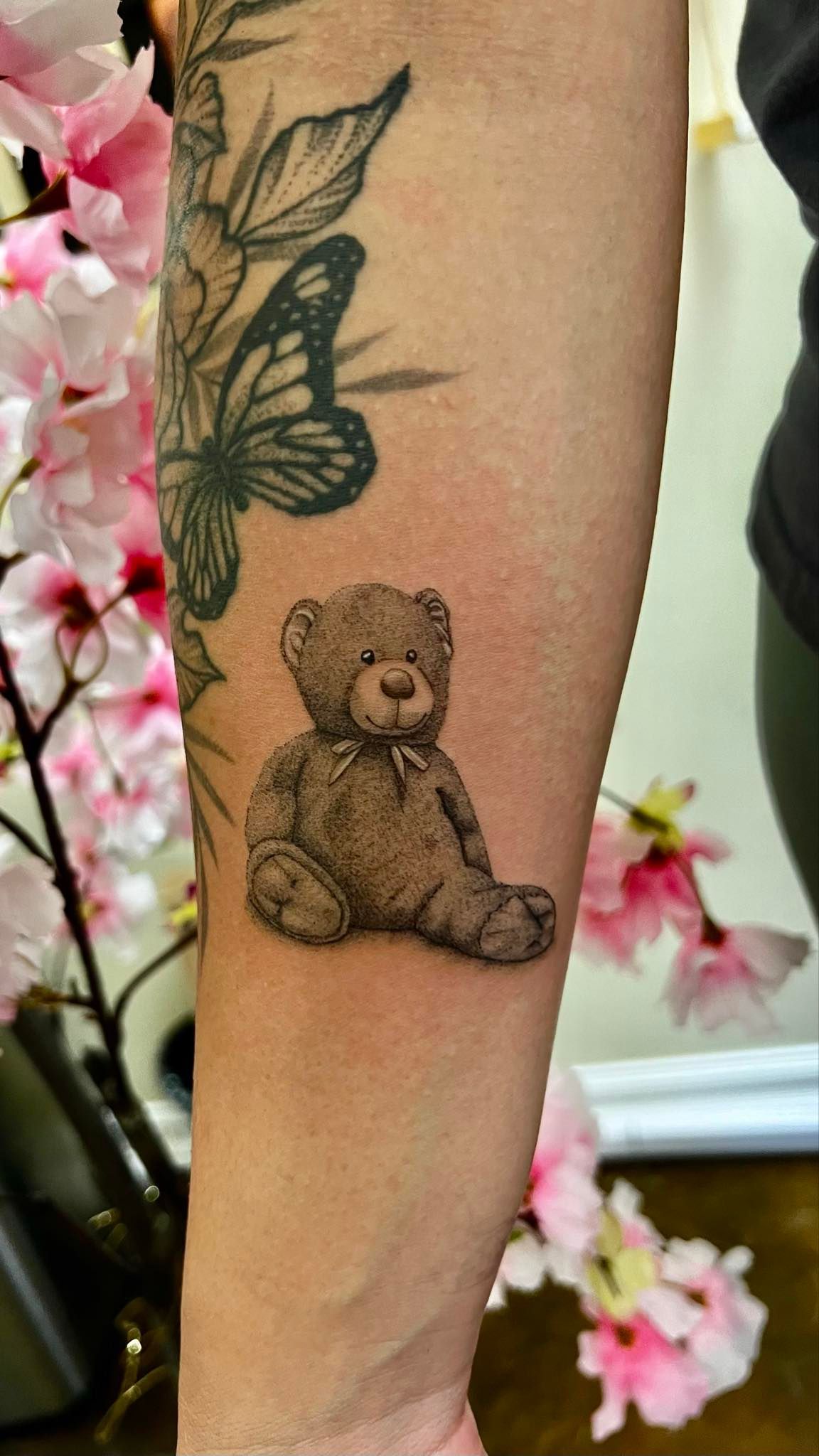 Tattoo of a seated brown teddy bear on an arm, with a butterfly tattoo and pink flowers in background.