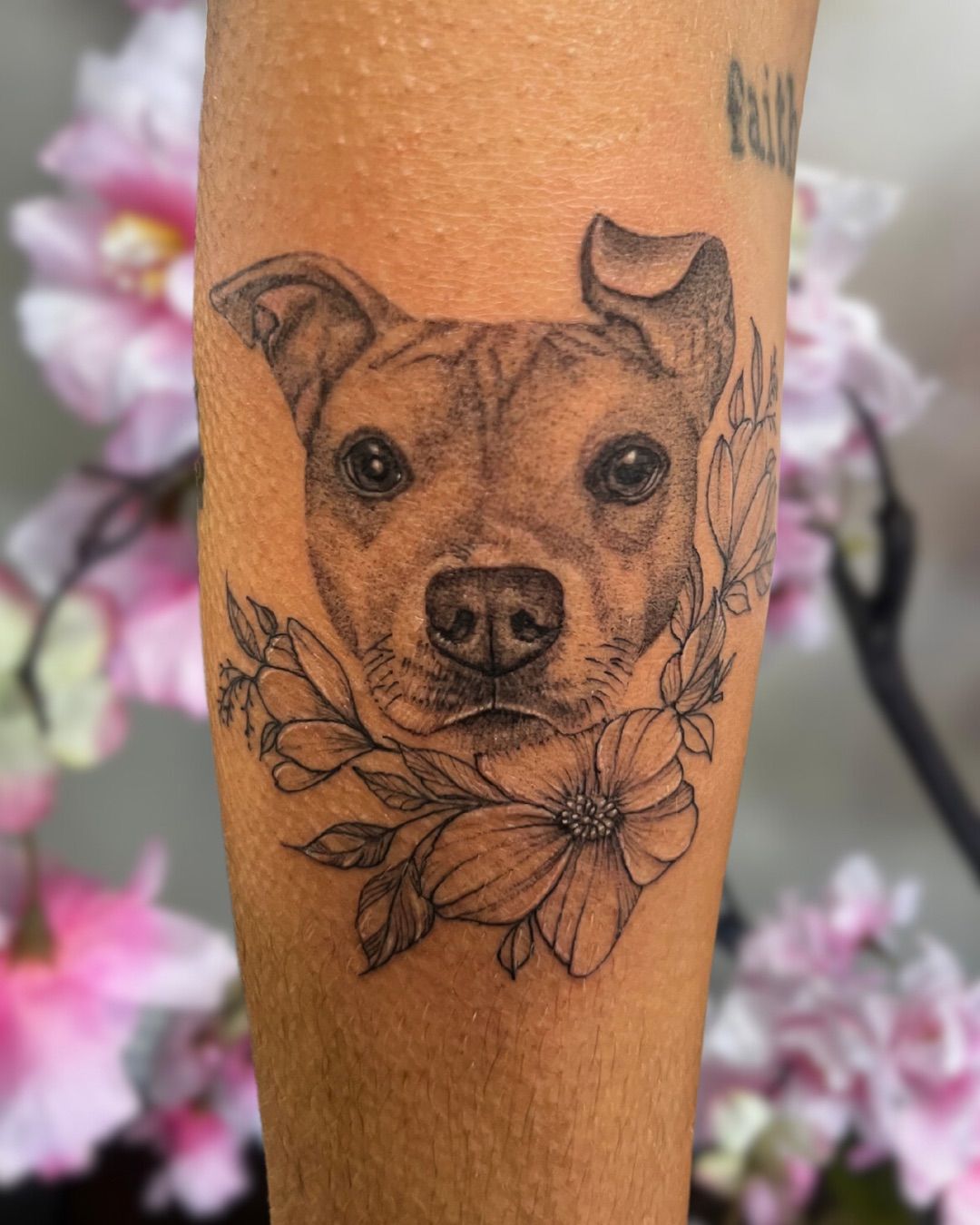 Tattoo of a dog's face surrounded by flowers on a person's arm. Black ink.