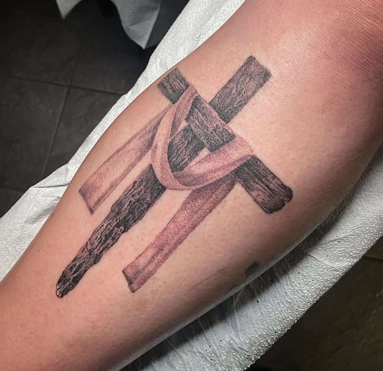 Tattoo of a black cross overlaid with a pink ribbon on a forearm.