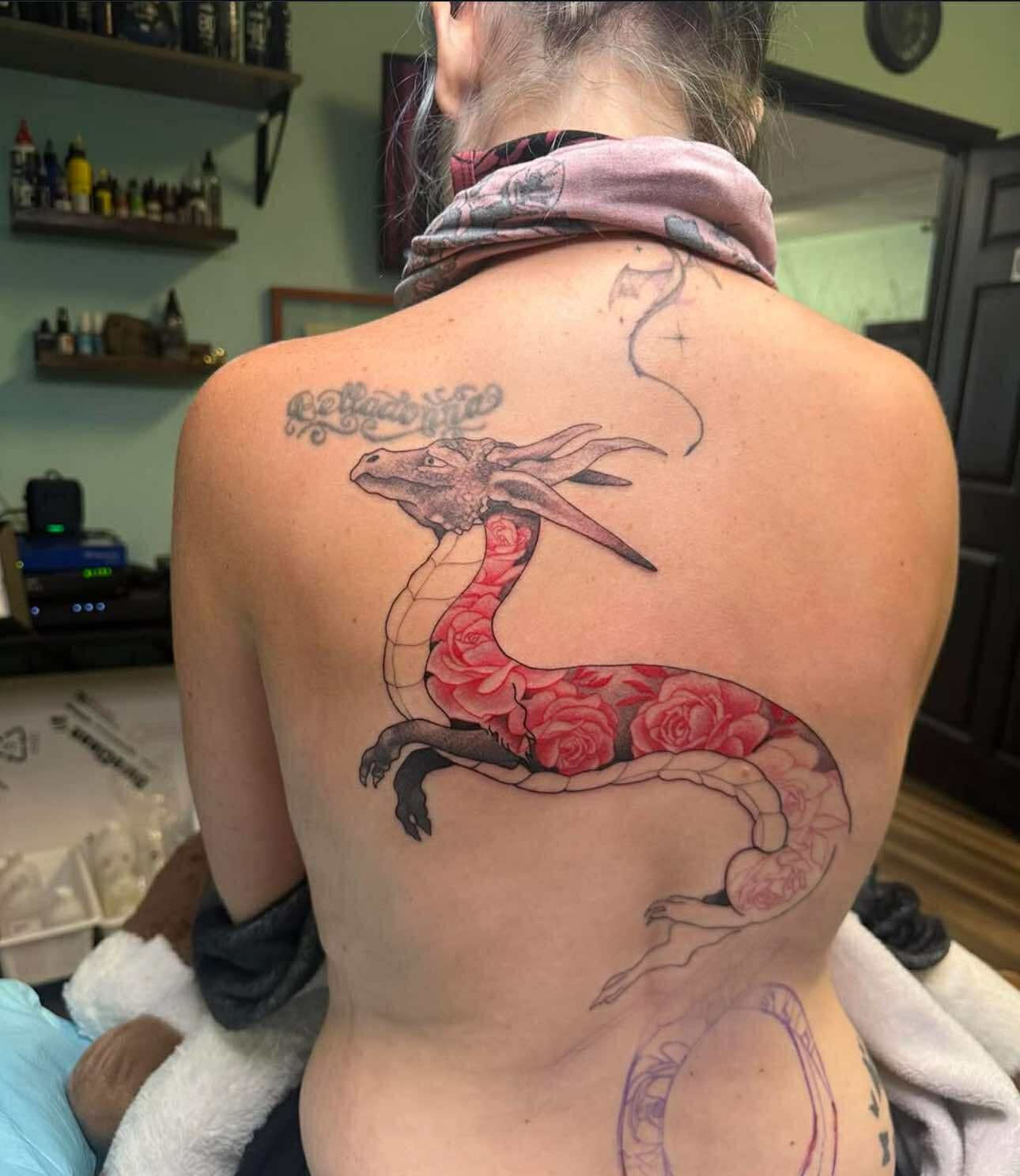 Woman's back with a dragon tattoo filled with red roses. The dragon is grey and black.