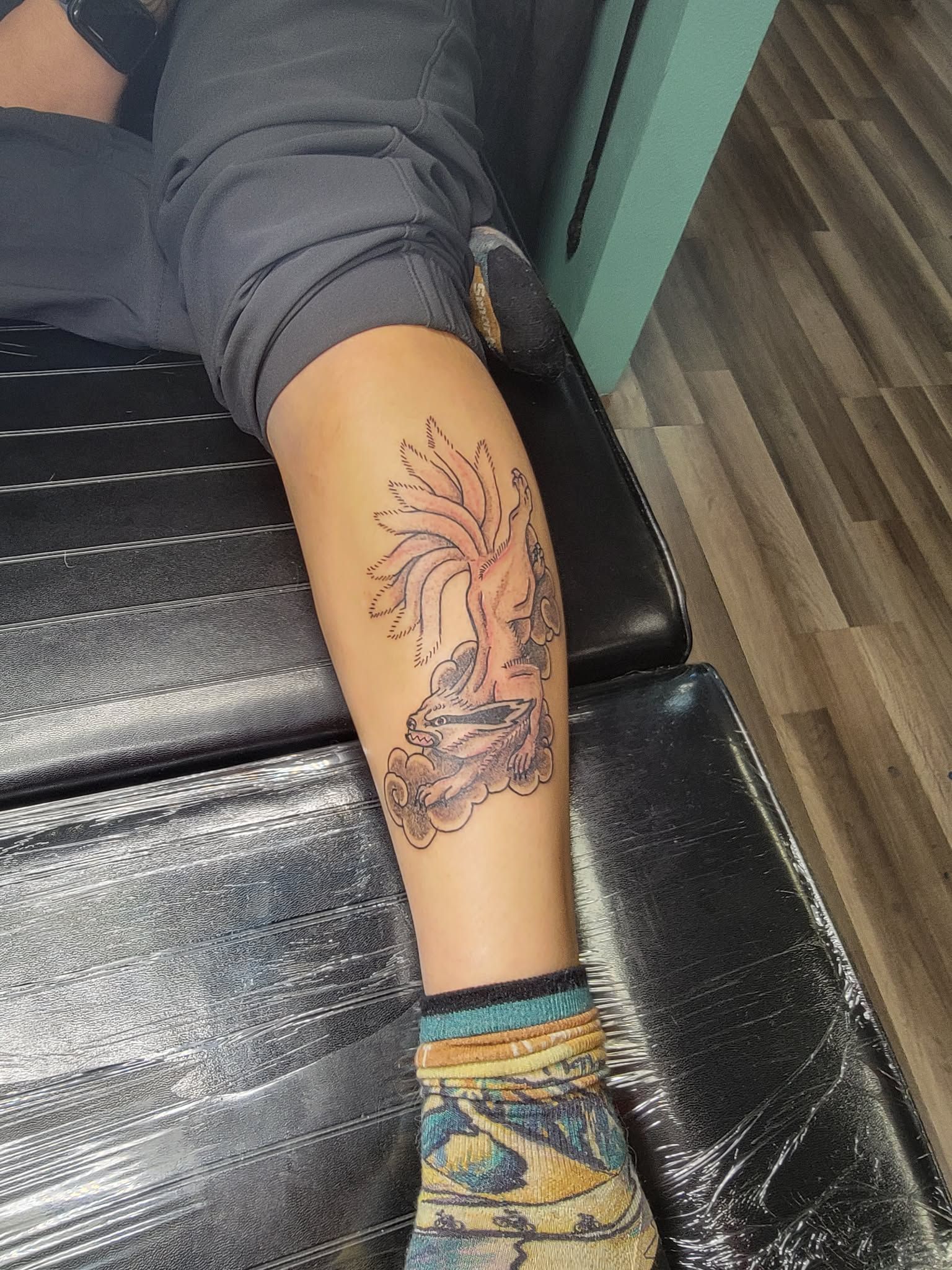 Leg with new tree tattoo, resting on a black surface. The person wears blue jeans and a patterned sock.
