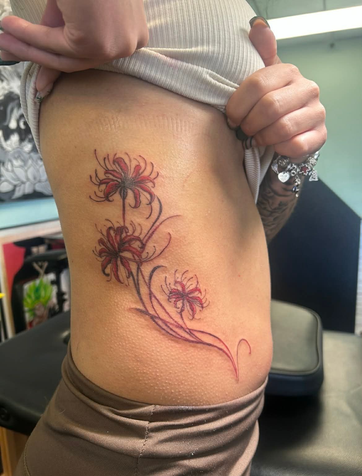 Tattoo of red spider lilies on a person's side, against their skin. The person wears a light shirt.