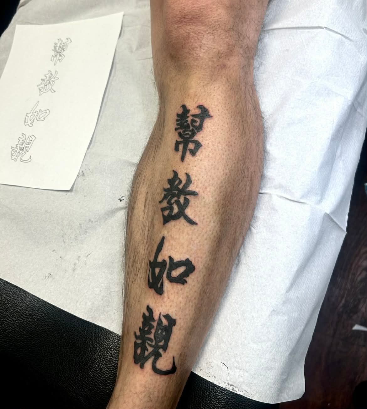 Black Chinese characters tattoo on a person's calf, next to a design sheet.