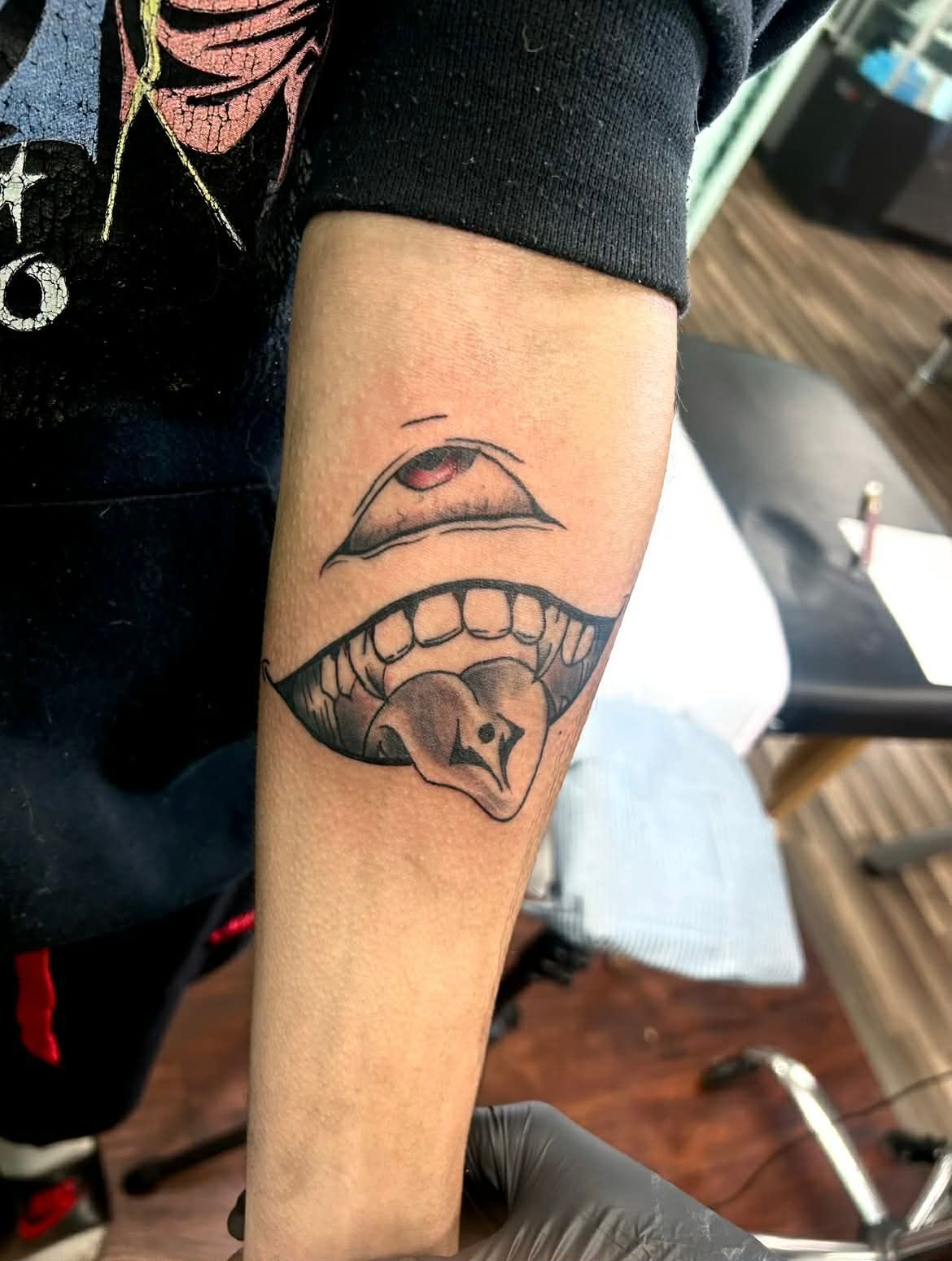 Arm tattoo of an open mouth with teeth and a protruding tongue, above an eye. Grayscale with some shading.